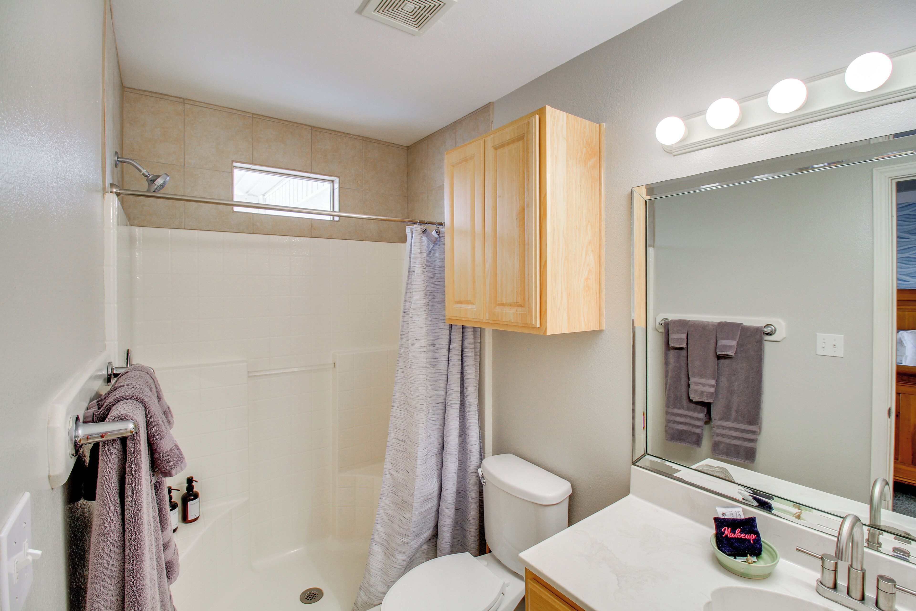A bathroom with a shower, toilet, vanity, and a mirror with lights.