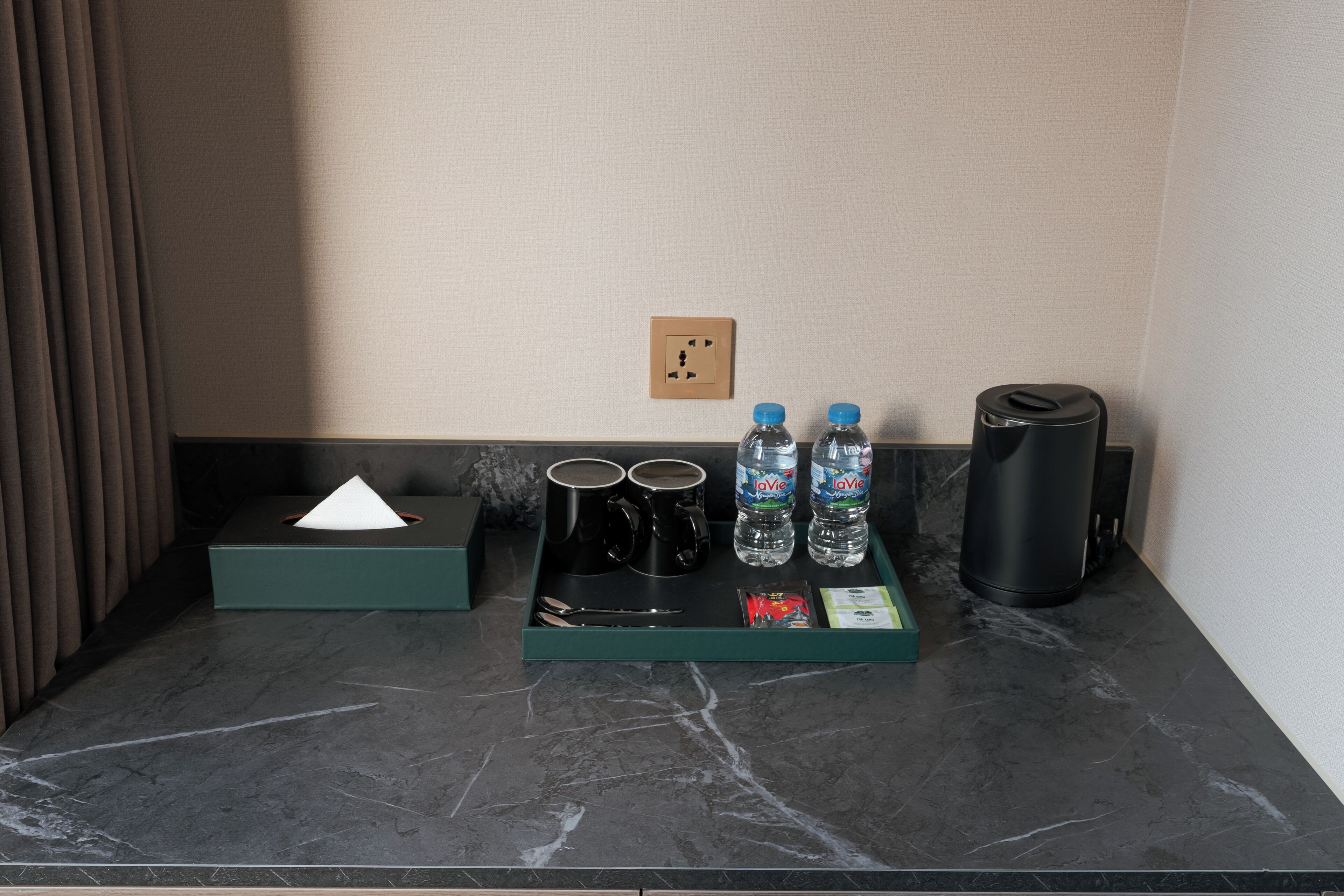 A hotel room's minibar with a tissue box, two black mugs, two LaVie water bottles, a black thermos, and a tray with a spoon and napkin.