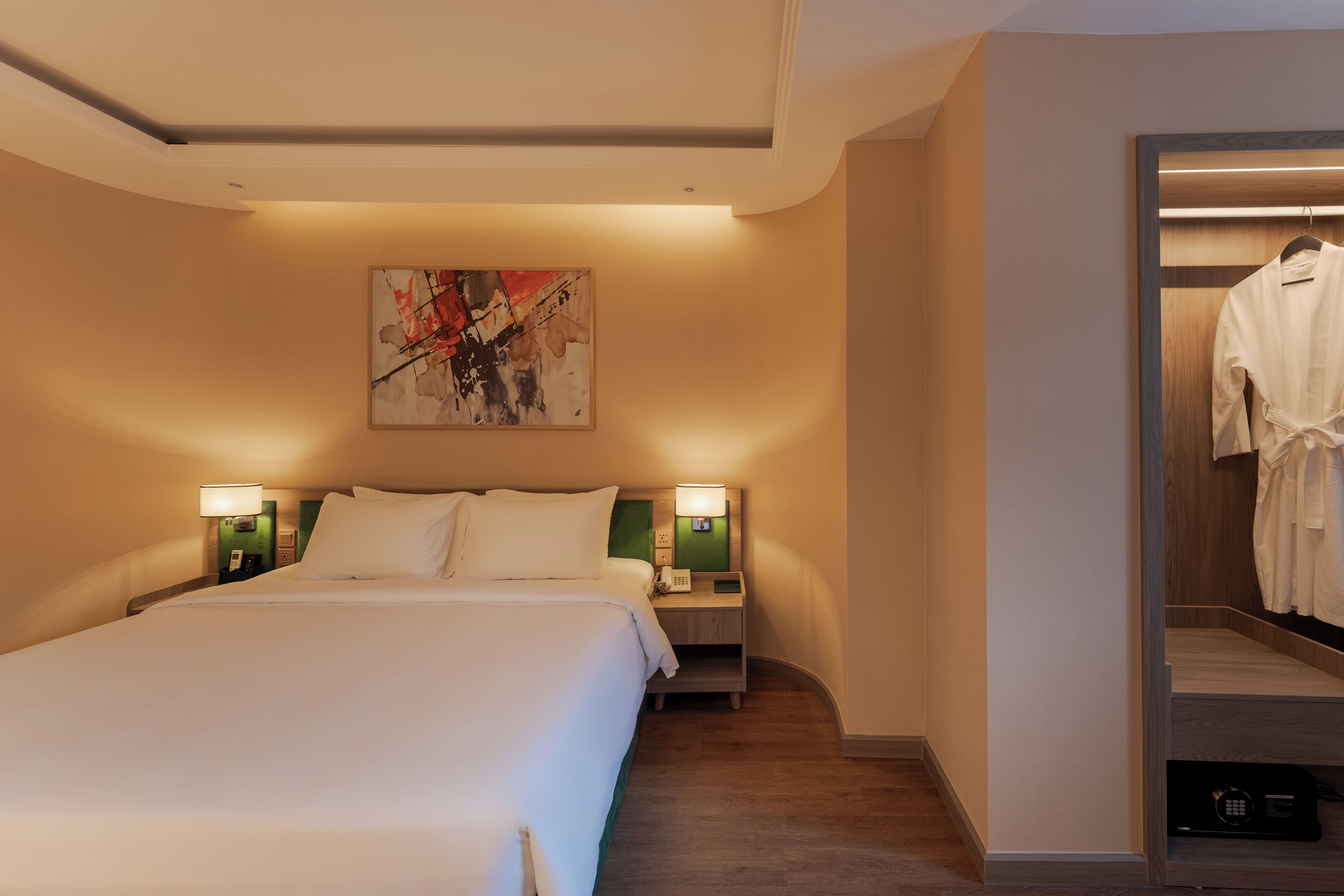 A hotel room with a large bed, two bedside tables with lamps, a painting on the wall, and a walk-in closet with a robe hanging.