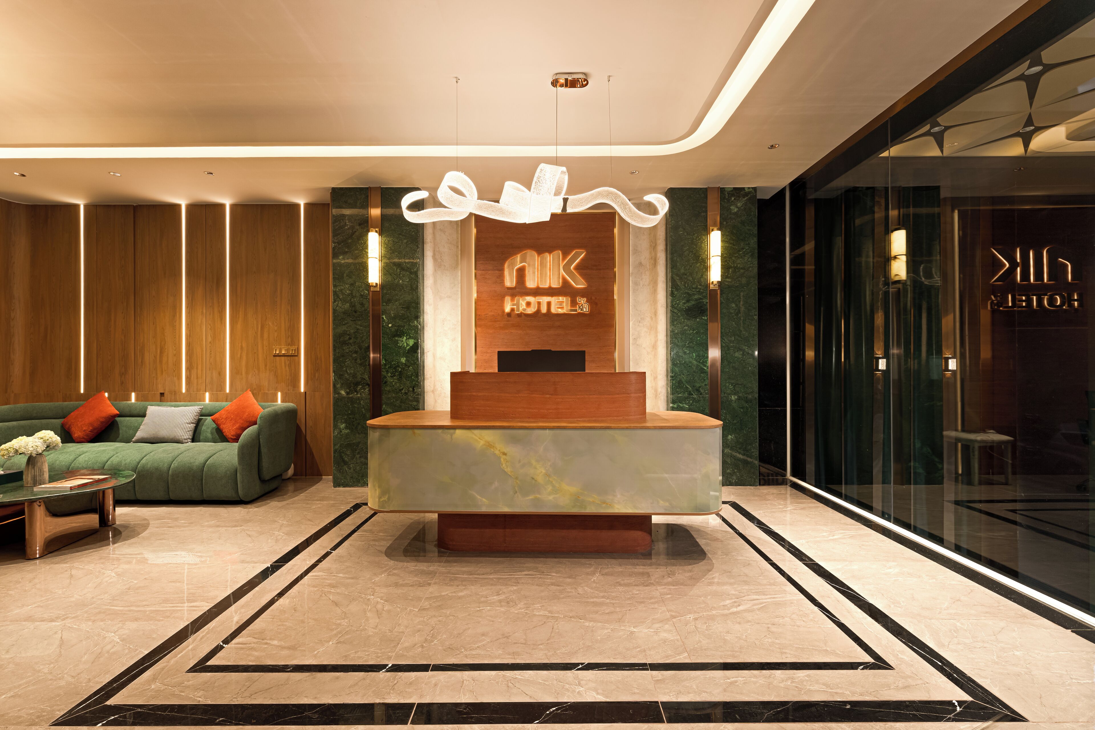 A modern hotel reception area with a marble reception desk, a green sofa with orange cushions, and a glass wall to the right.