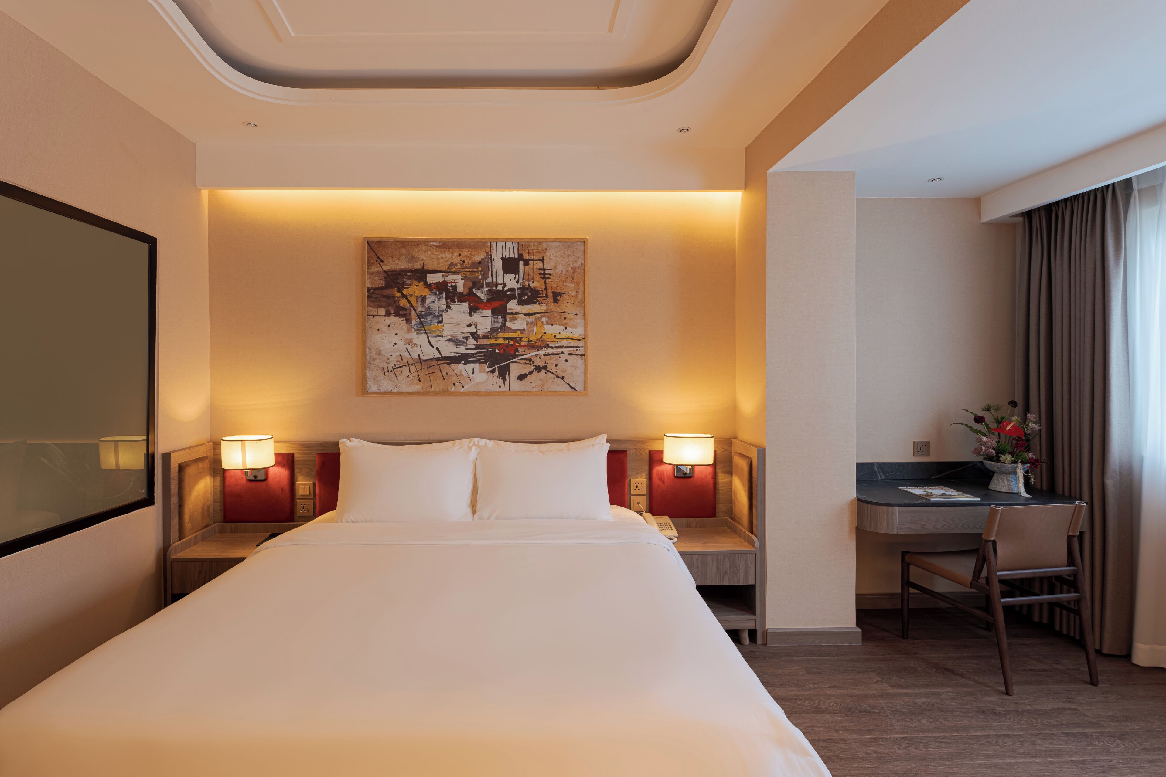 Quarto de hotel com uma cama grande, duas luminárias de cabeceira, uma escrivaninha com cadeira e um quadro na parede.