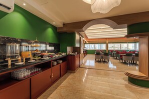 Un comedor moderno de hotel con barra libre, una zona de estar con mesas y sillas, y vistas a la ciudad a través de amplias ventanas.