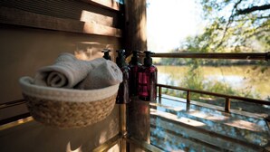 A wooden cabin with a glass railing overlooking a lake. A woven basket with towels and toiletries is visible on a table.