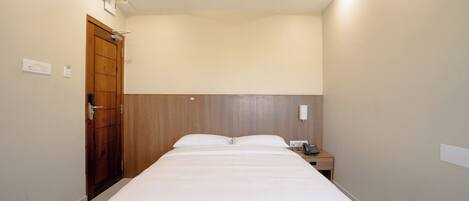 Una camera d'albergo con un letto singolo, una testiera in legno, un comodino con un telefono e una porta con maniglia.