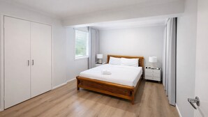 Een slaapkamer met een houten bed, witte gordijnen, een nachtkastje met een lamp en een kledingkast.