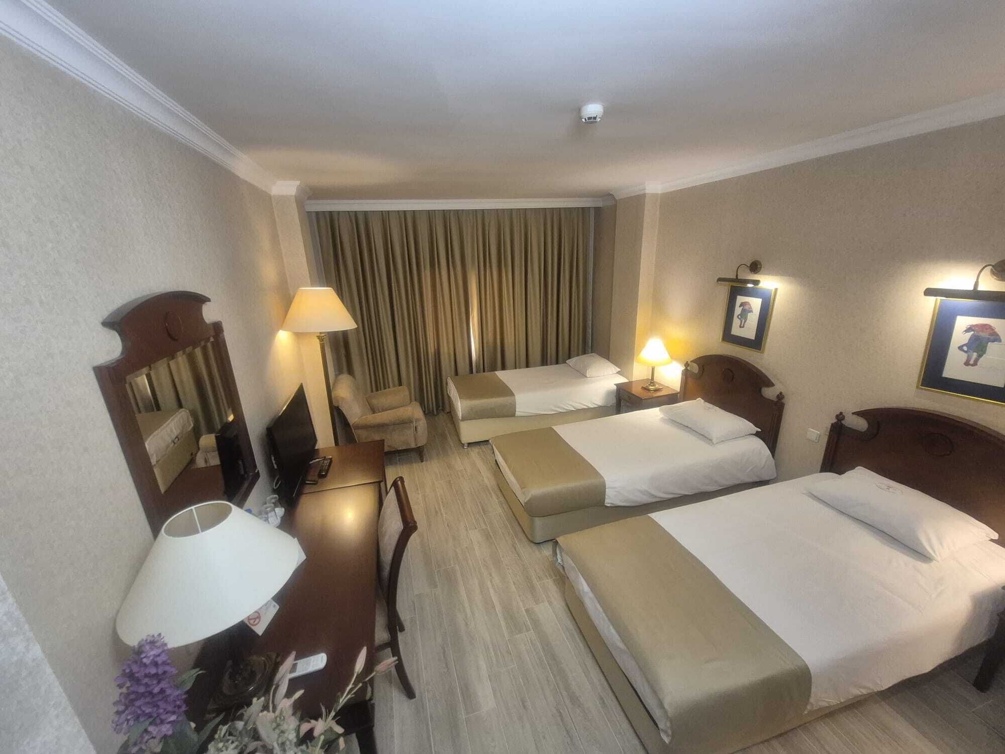 A hotel room with two beds, a desk, a chair, a lamp, and a large window with curtains.