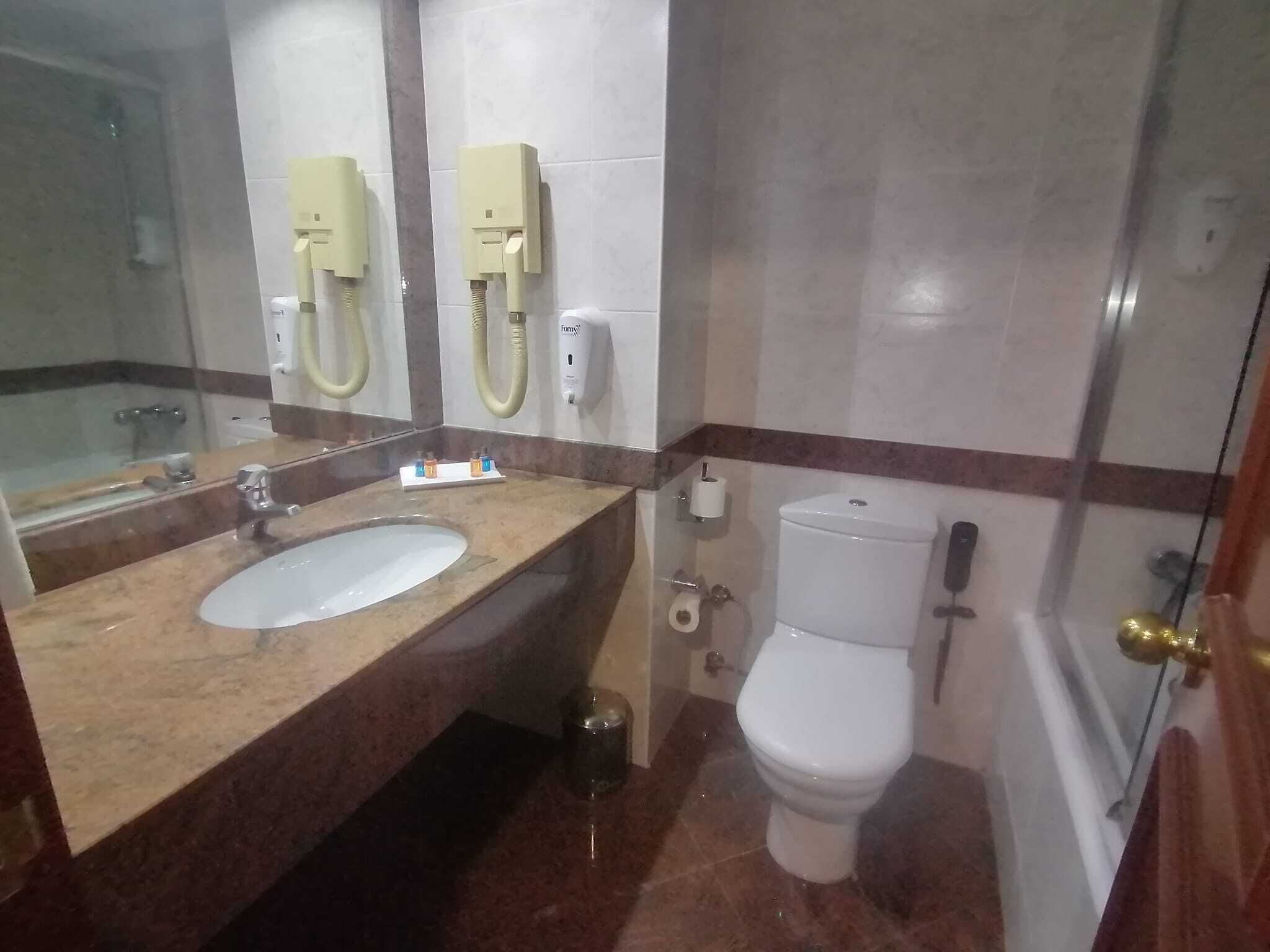 Une salle de bain avec un lavabo en marbre, deux sèche-mains, des toilettes et une poubelle.