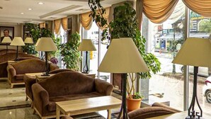 Um saguão de hotel com várias áreas de estar, incluindo sofás e poltronas, dispostas em torno de uma mesa de centro. Há diversas plantas em vasos e uma escada que leva a um andar superior.