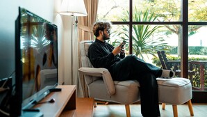 Una persona sentada en un sofá en una habitación con televisión, una lámpara y vistas a áreas verdes al exterior.