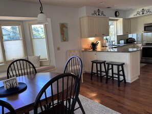 A kitchen with white cabinets, a central island with stools, a dining area with a table and chairs, and a window with blinds. - FairView: Sea Pines Retreat: golf views, screen porch, pool, privacy. STR#077244 (Hilton Head Island)