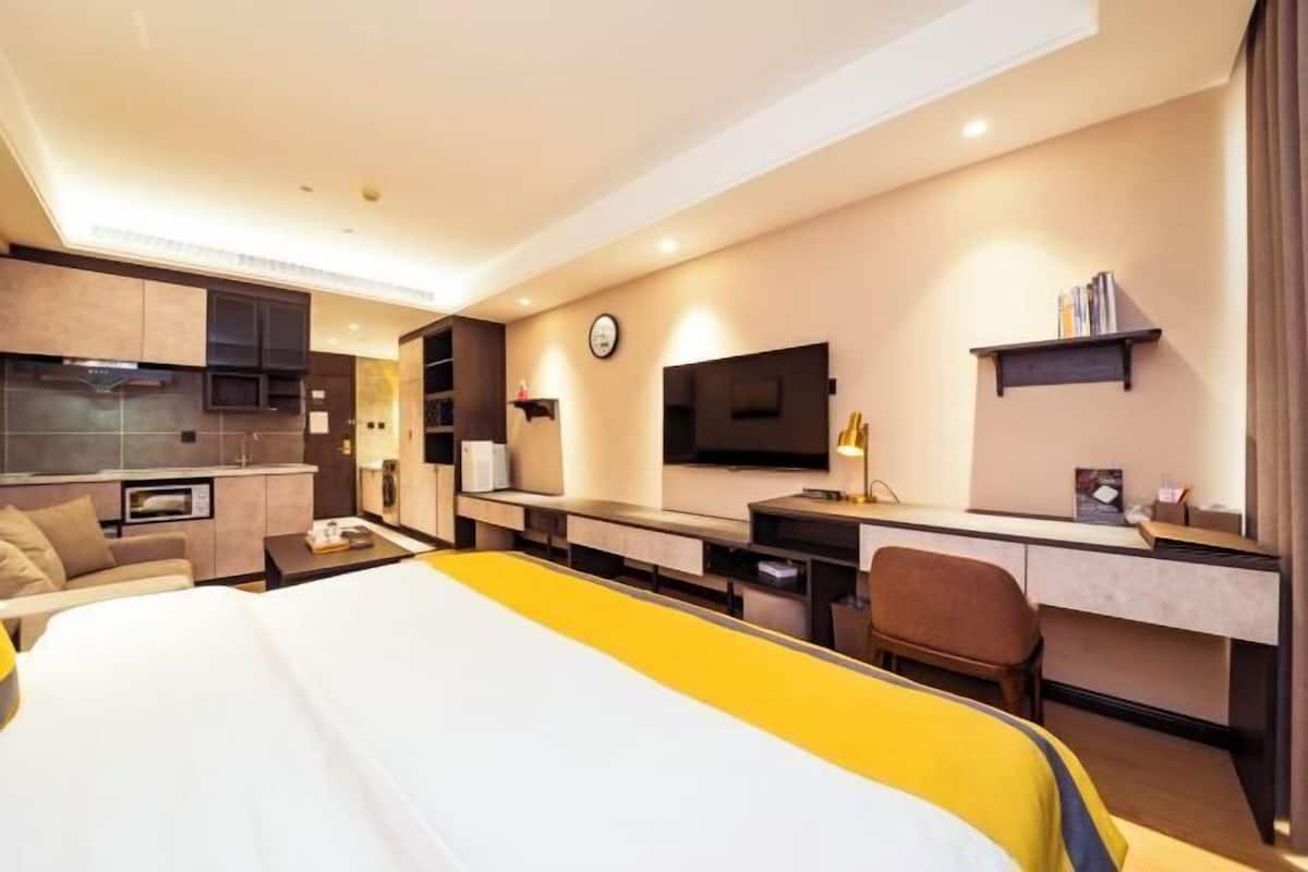 A modern hotel room with a bed, a desk, a chair, a TV, a clock, and a small kitchenette.