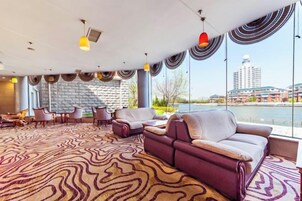 Amplia sala de estar con alfombra estampada, diferentes disposiciones de asientos y grandes ventanales que ofrecen vistas a un cuerpo de agua y edificios.