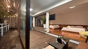 Quarto de hotel com piso de madeira, uma cama, televisão fixada na parede e uma pequena mesa com um vaso de flores.
