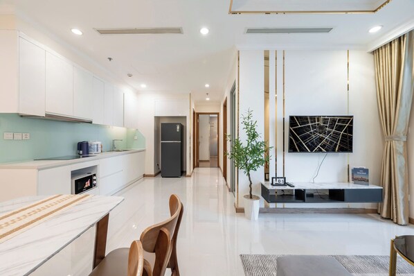 A modern kitchen with white cabinets, a built-in microwave, and a dining area with a marble table and wooden chairs. - Vinhomes Urban Apartment HCM (Ho Chi Minh City)
