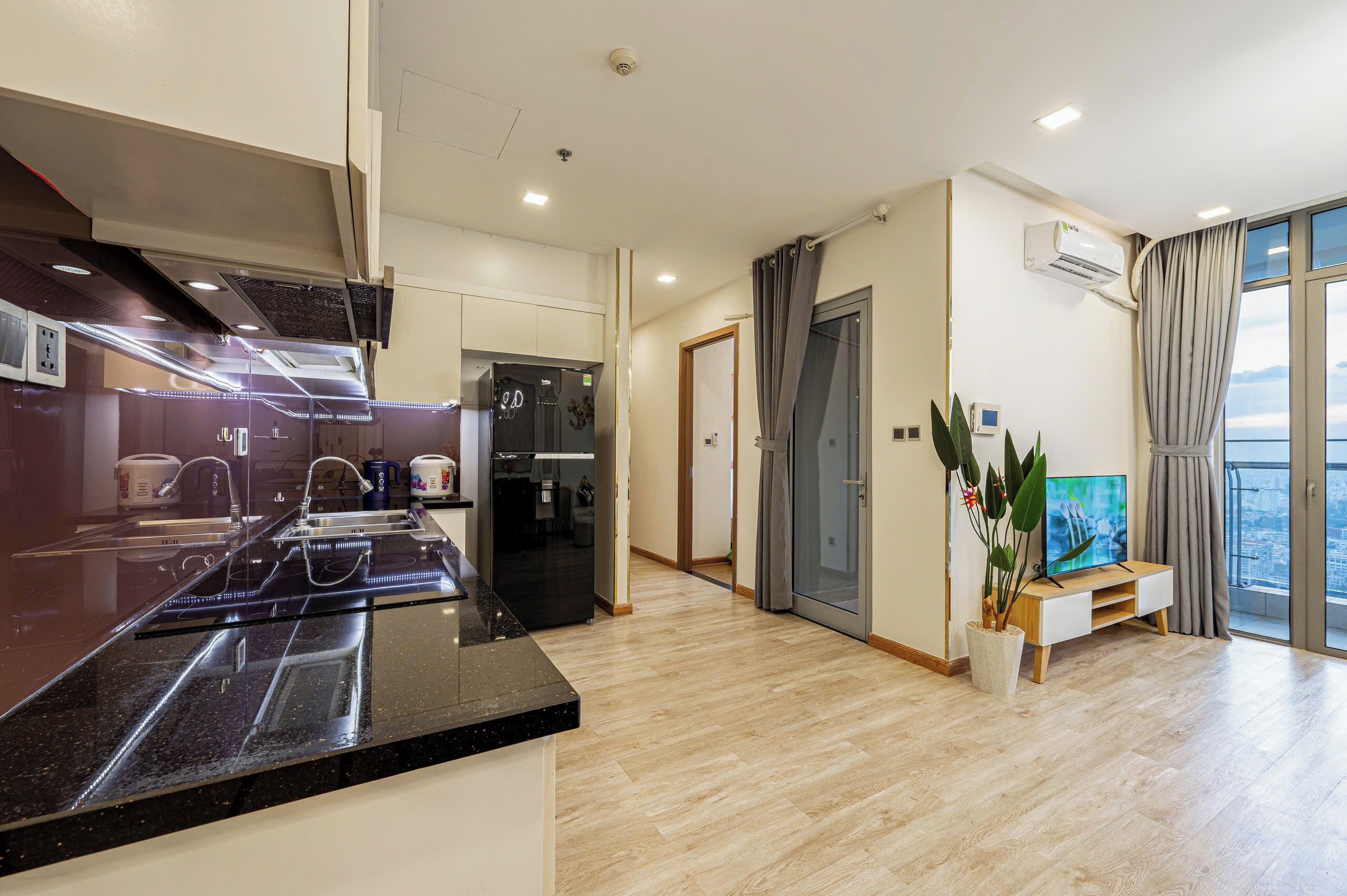 Een moderne keuken met een zwart aanrecht, een inbouwoven en een koelkast. Er staat een houten eettafel met stoelen, een potplant en een flatscreen-tv op een houten standaard.