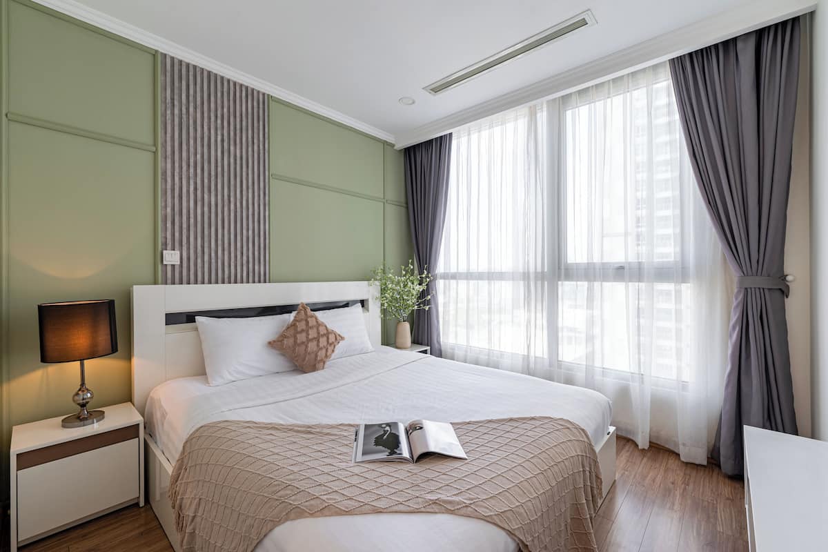 Ein ordentlich eingerichtetes Hotelzimmer mit einem großen Bett, einem Nachttisch mit Lampe und einem Fenster mit Vorhängen.