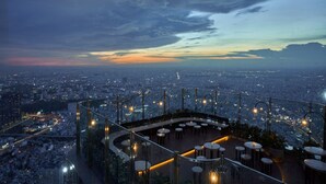 Un bar en la azotea con vistas panorámicas de la ciudad, barandillas de vidrio y mesas redondas.