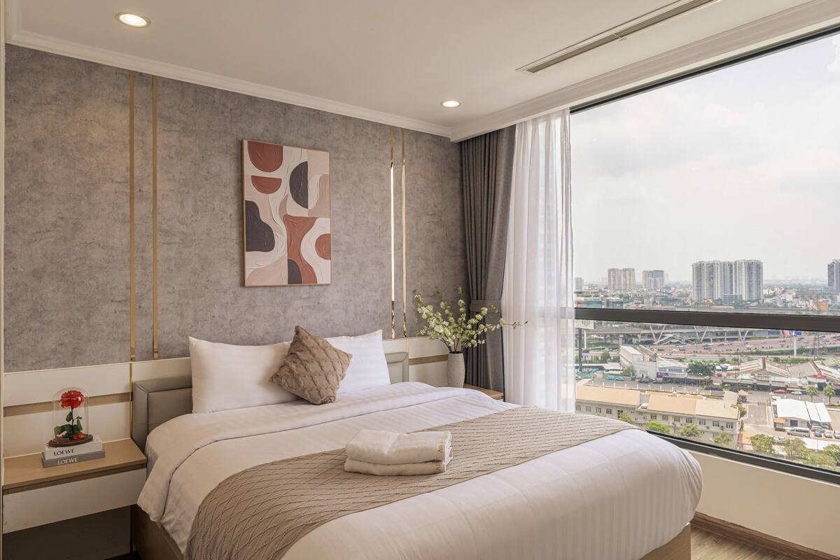 Ein Hotelzimmer mit einem großen Bett, Blick auf die Stadt, einem Bild an der Wand und einer Vase mit Blumen.