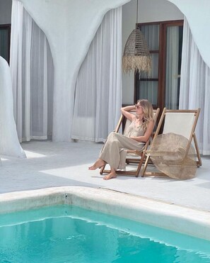 A person sitting on a wooden chair by a pool with a hanging light fixture. - Sal Central (Pecatu)