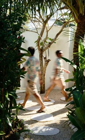 Two people walking on a pathway surrounded by greenery and a white wall. - Sal Central (Pecatu)
