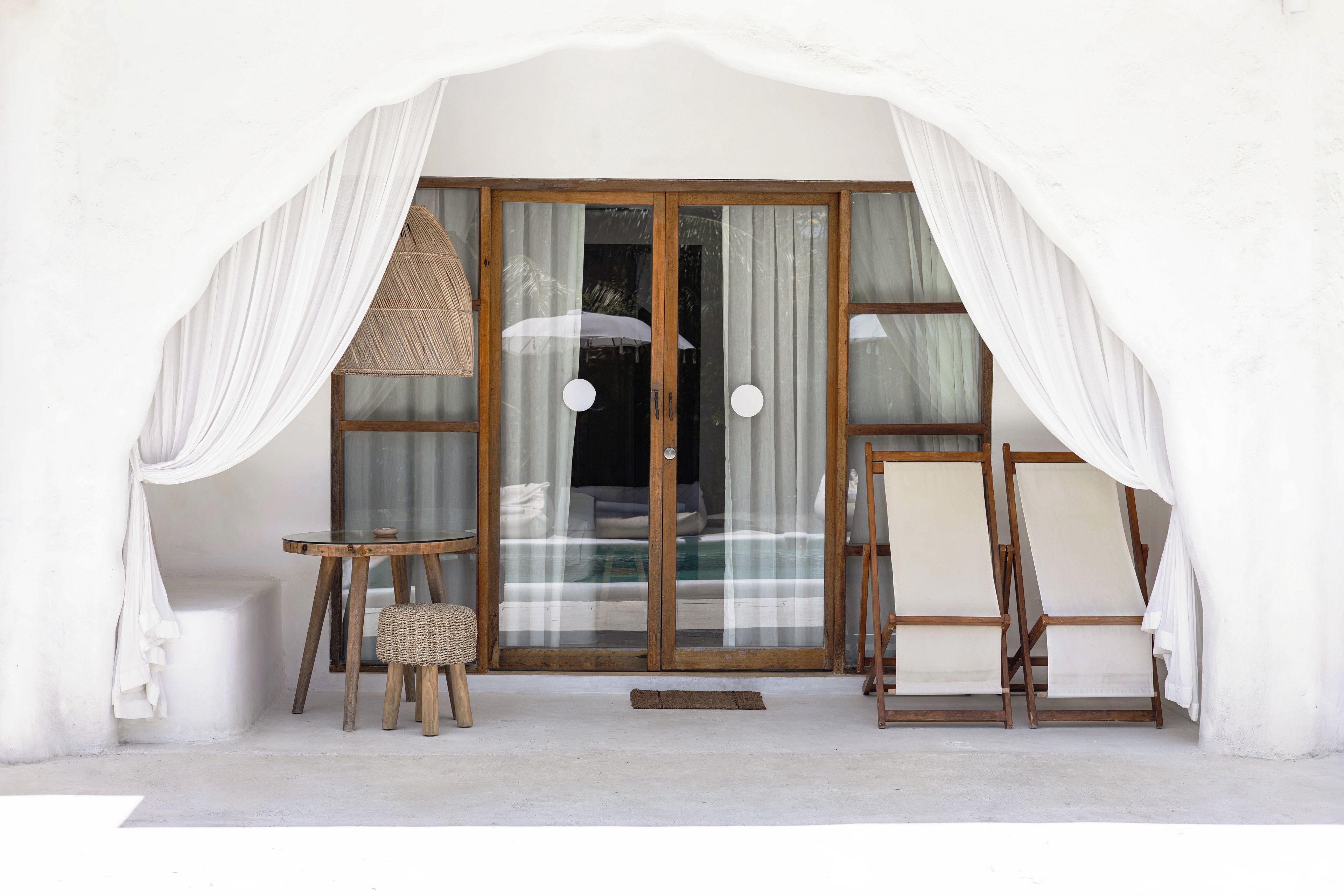 Una habitación con puerta de cristal, cortinas blancas, una silla de mimbre, una mesita redonda y una silla de madera.