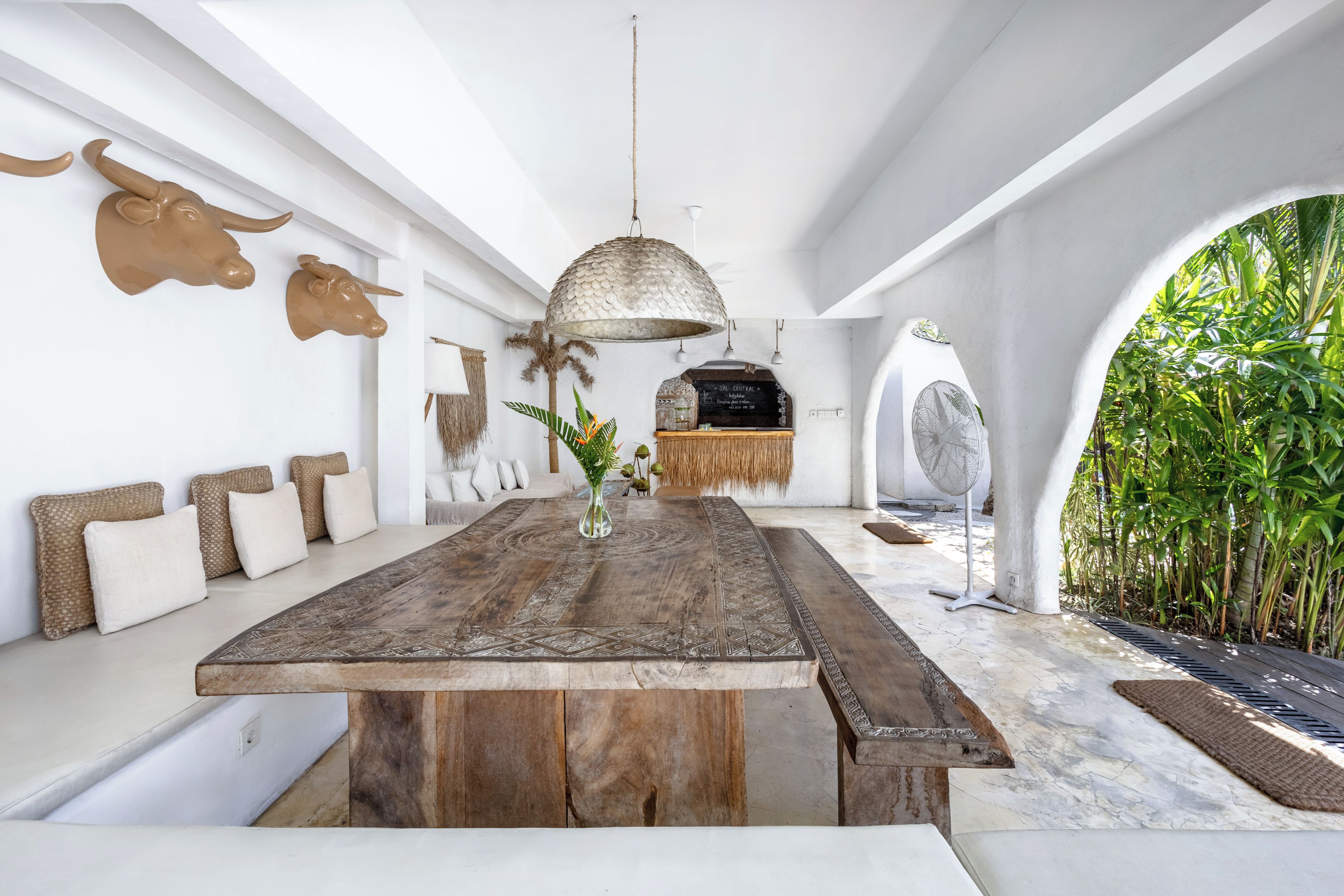 A spacious room with a long wooden table, a hanging light fixture, and a view of a lush garden through an archway.