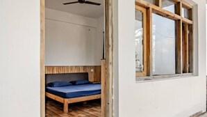 Een kamer met een houten vloer, een bed met een blauwe matras, een plafondventilator en een raam met houten kozijnen.