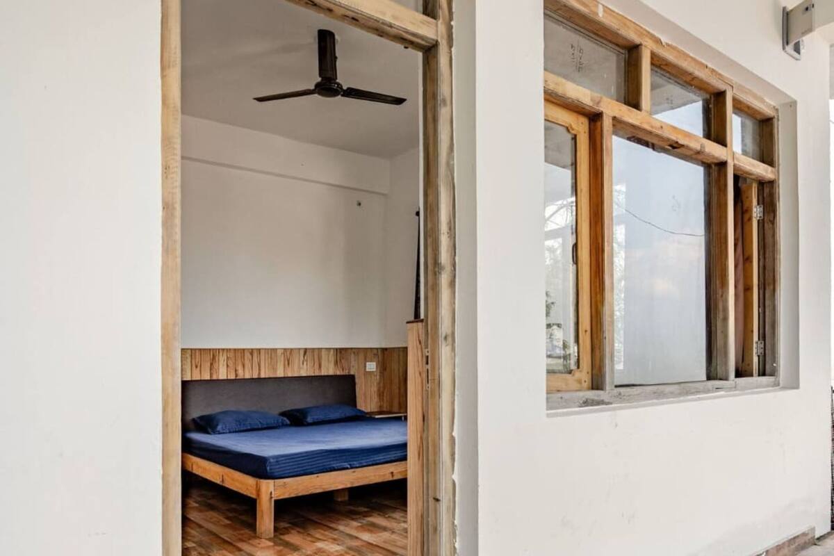 A room with a wooden floor, a bed with a blue mattress, a ceiling fan, and a window with wooden frames.