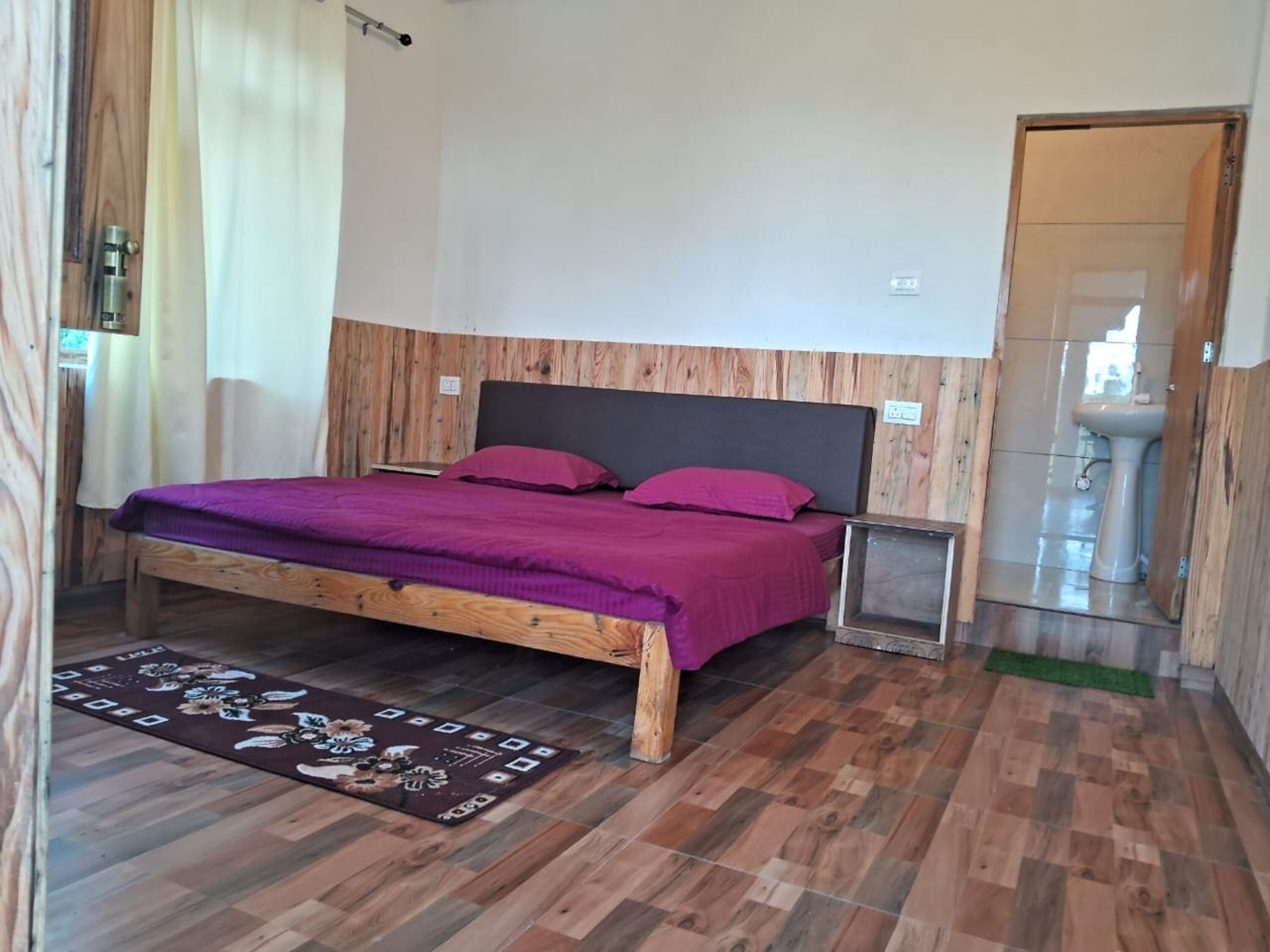 Una habitación con una cama de madera, una mesita de noche pequeña, un baño con lavamanos y una alfombra estampada.