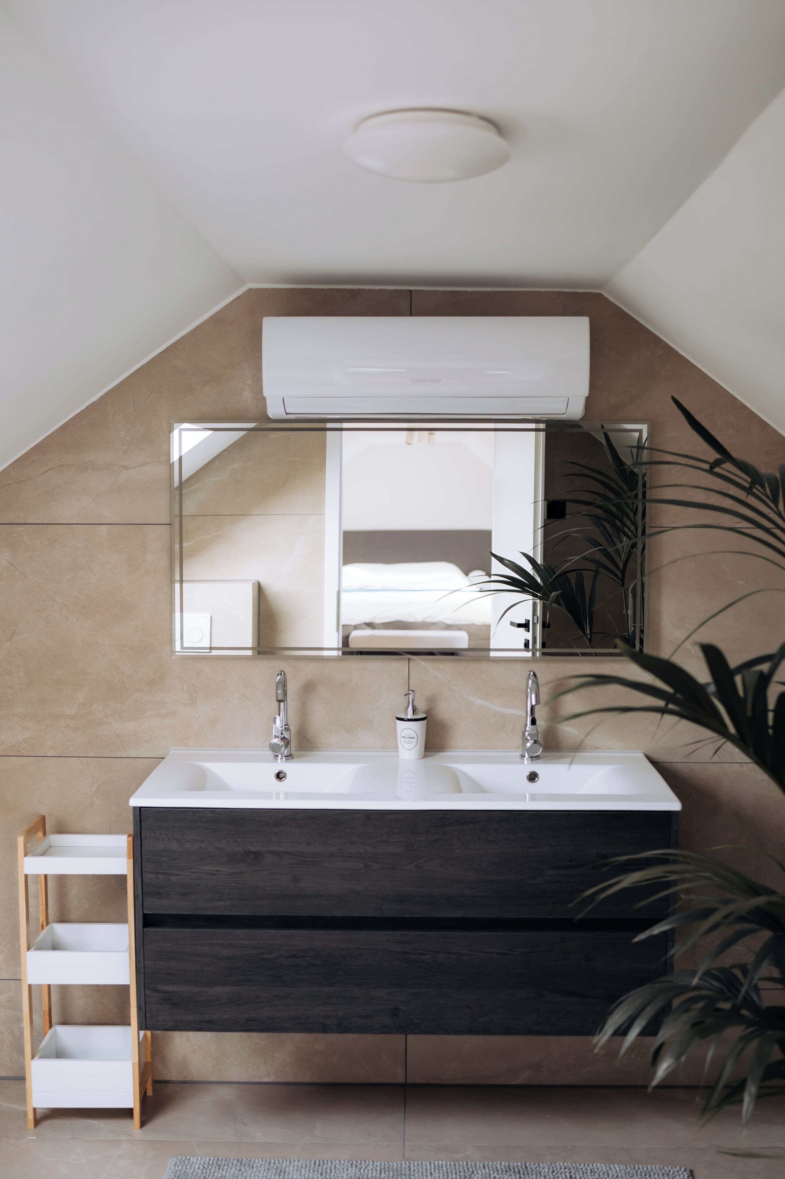 A modern bathroom with a large mirror, two sinks, a dark wooden vanity, a white shelf, and a ceiling-mounted air conditioner.