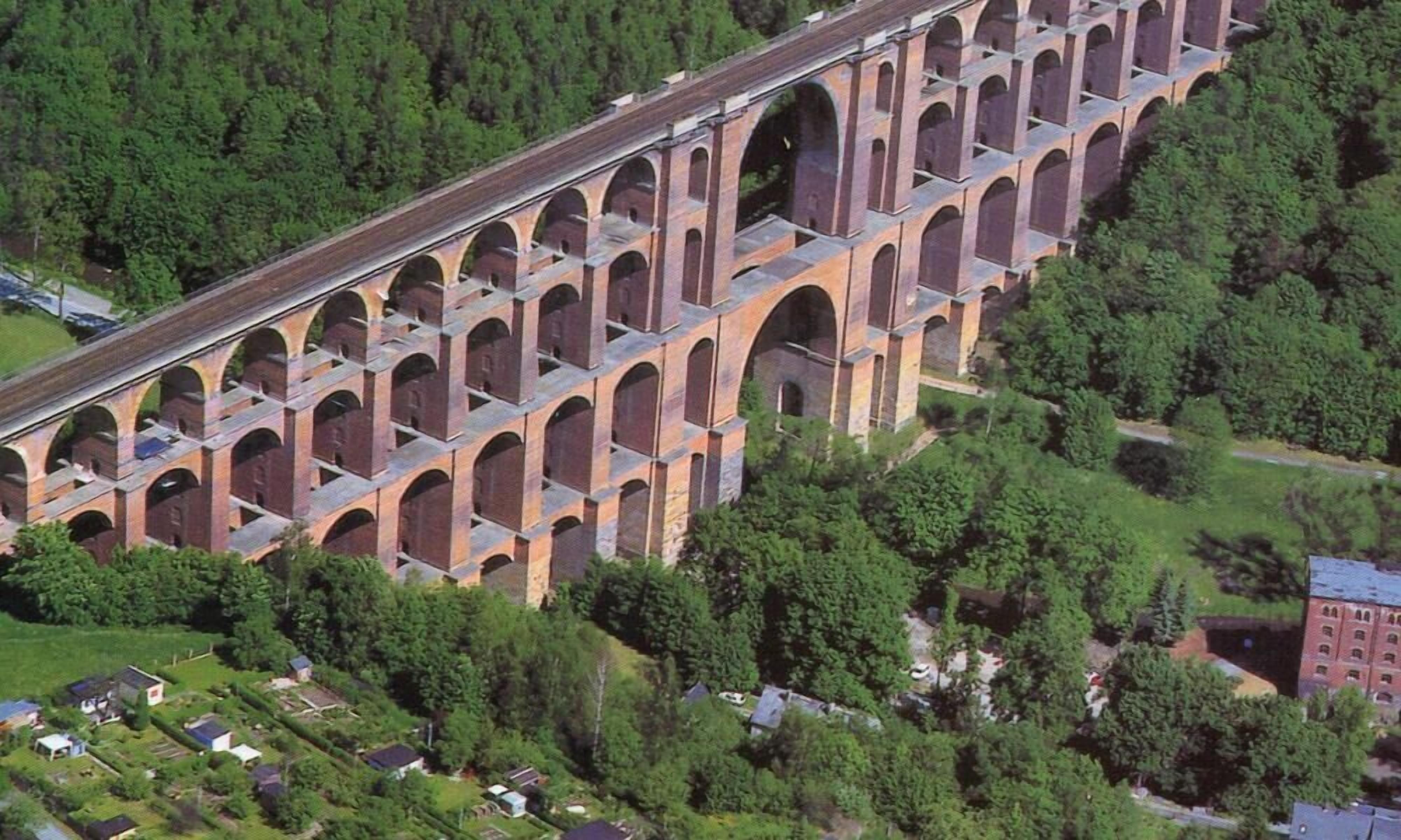 Un grand viaduc en briques, doté de nombreuses arches enjambant une vallée, est entouré d’une végétation luxuriante et de zones résidentielles.