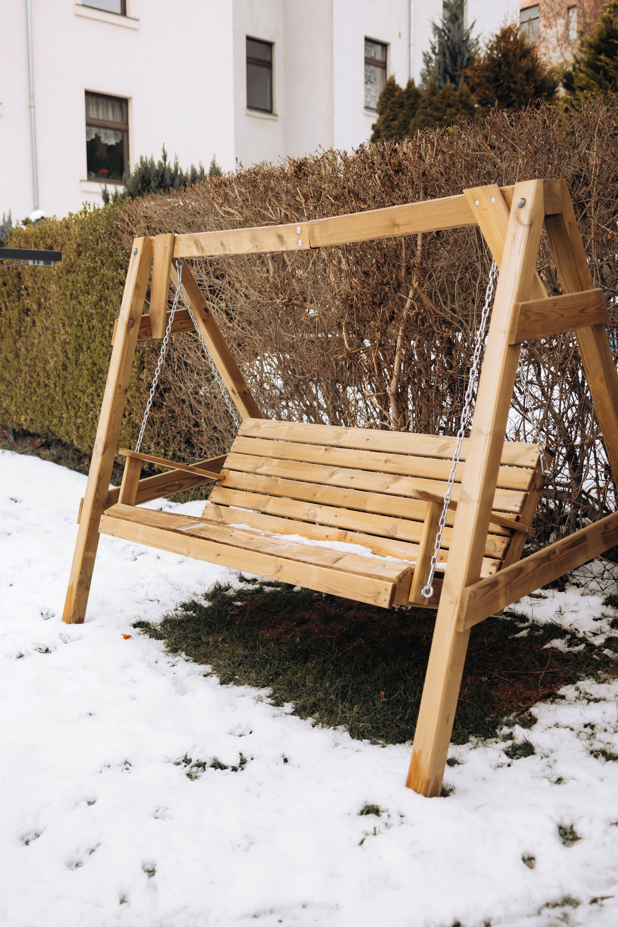 A wooden swing set in a snowy outdoor area with a hedge and a building in the background.