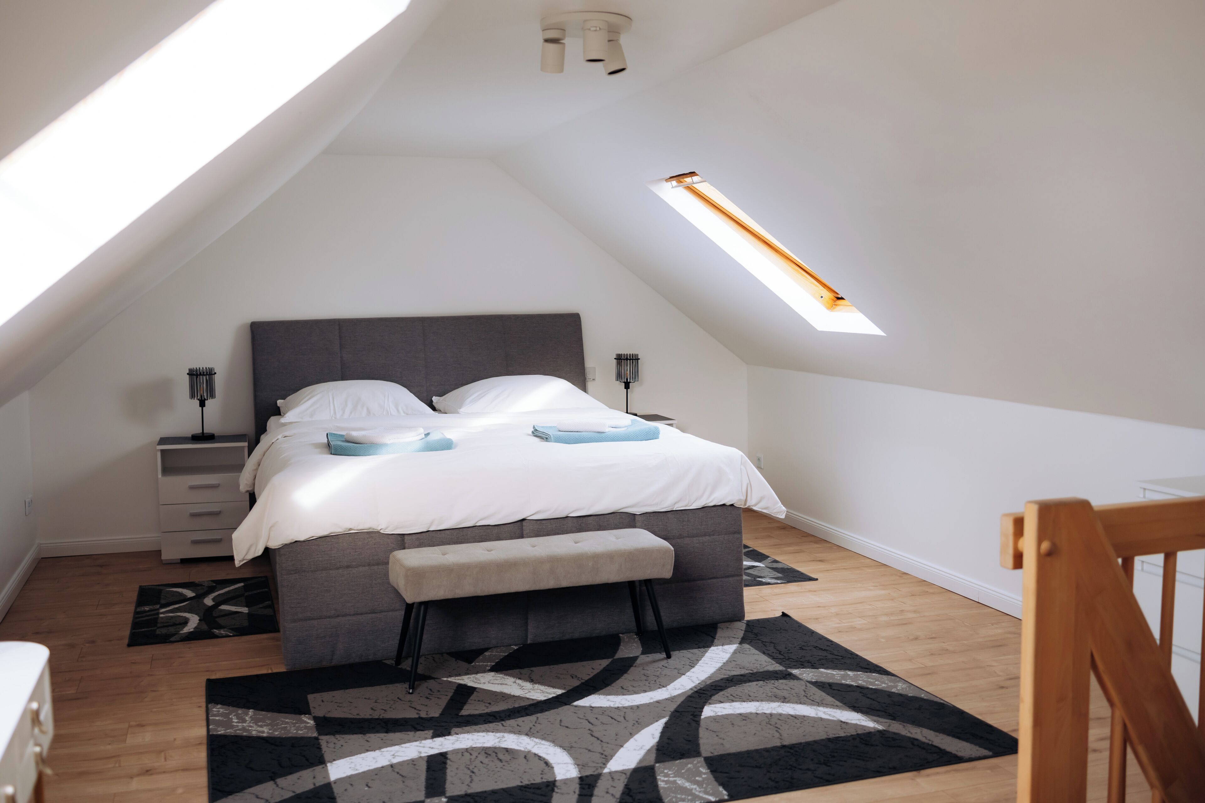 A bedroom with a bed, bedside tables, a bench, a skylight, and a wooden floor.