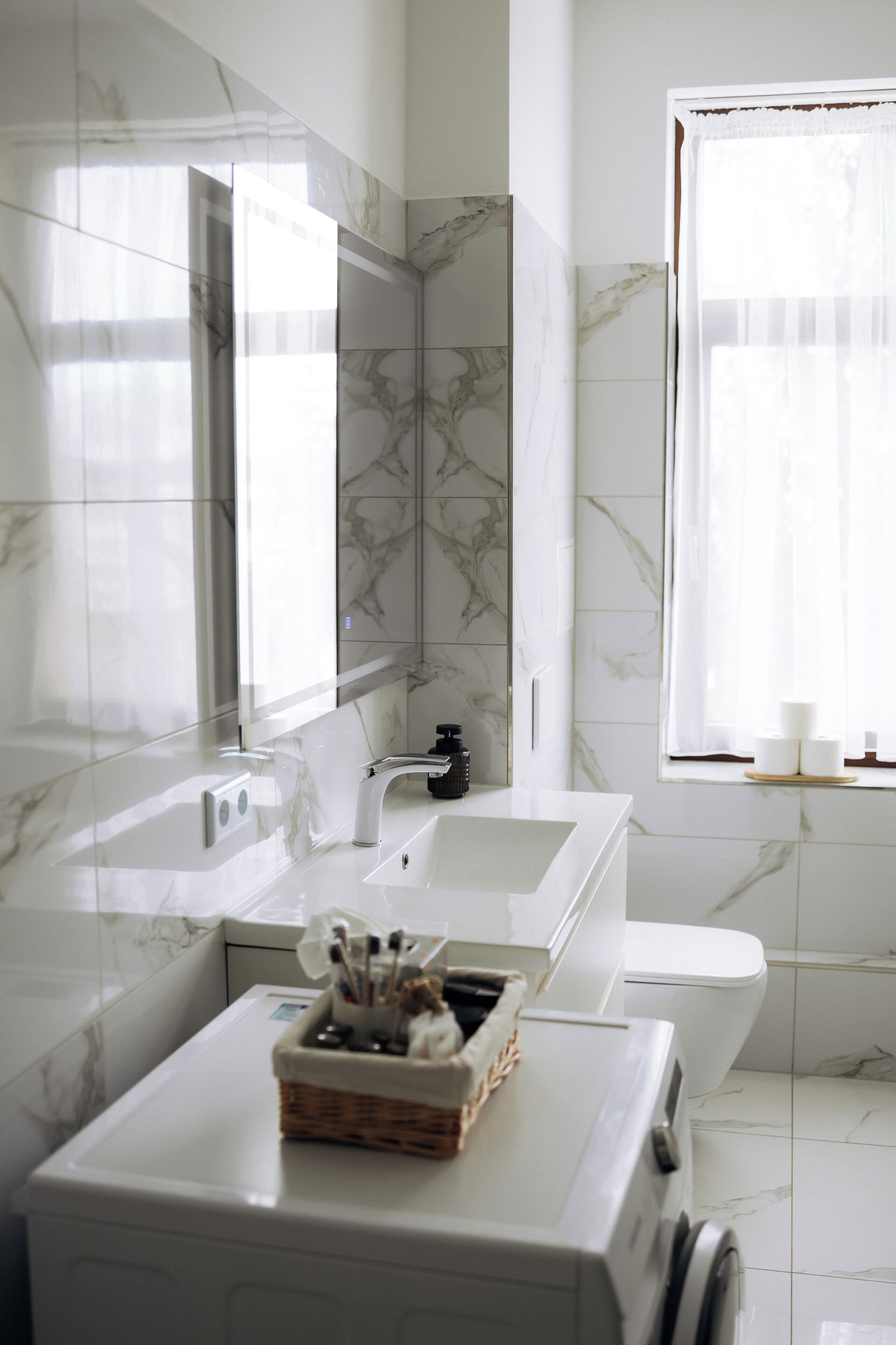 A modern bathroom with a marble backsplash, a large mirror, a white sink, a toilet, and a window with sheer curtains.