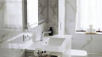 A modern bathroom with a marble backsplash, a large mirror, a white sink, a toilet, and a window with sheer curtains.