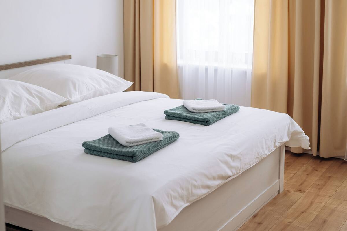 Una cama bien hecha con sábanas blancas y dos toallas dobladas, una lámpara de noche y una ventana con cortinas beige.