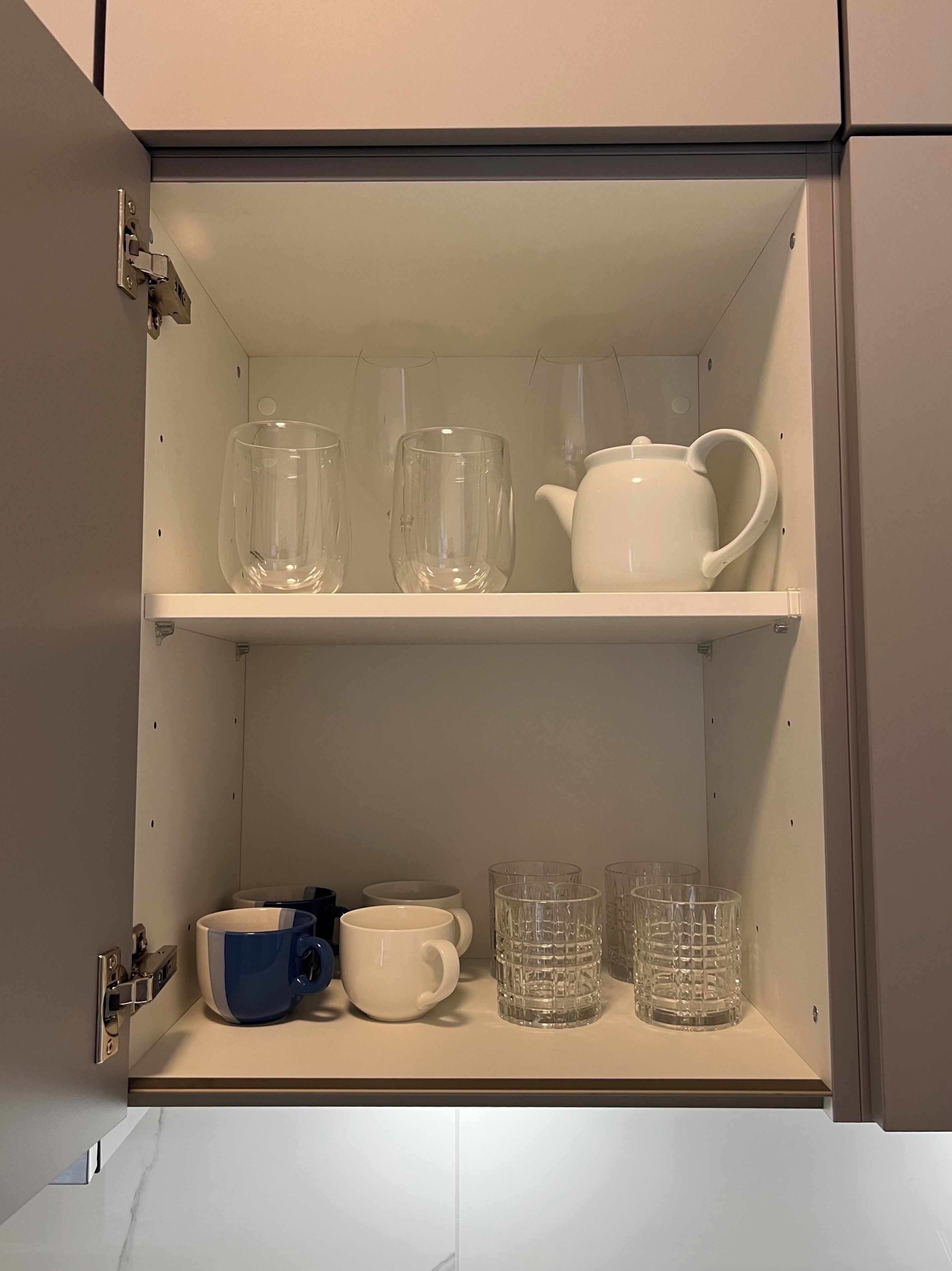 Un mueble de cocina con dos repisas, una con cristalería transparente y la otra con una tetera blanca y tazas.