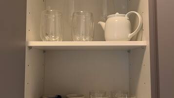 Un mueble de cocina con dos repisas, una con cristalería transparente y la otra con una tetera blanca y tazas.