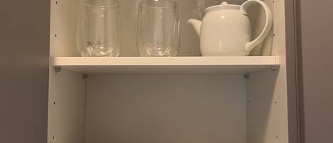 A kitchen cabinet with two shelves, one holding clear glassware and the other holding a white teapot and cups.