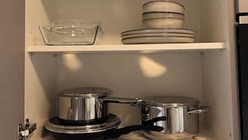Un meuble de cuisine contenant un récipient en verre transparent, des assiettes empilées et des ustensiles de cuisine en acier inoxydable.