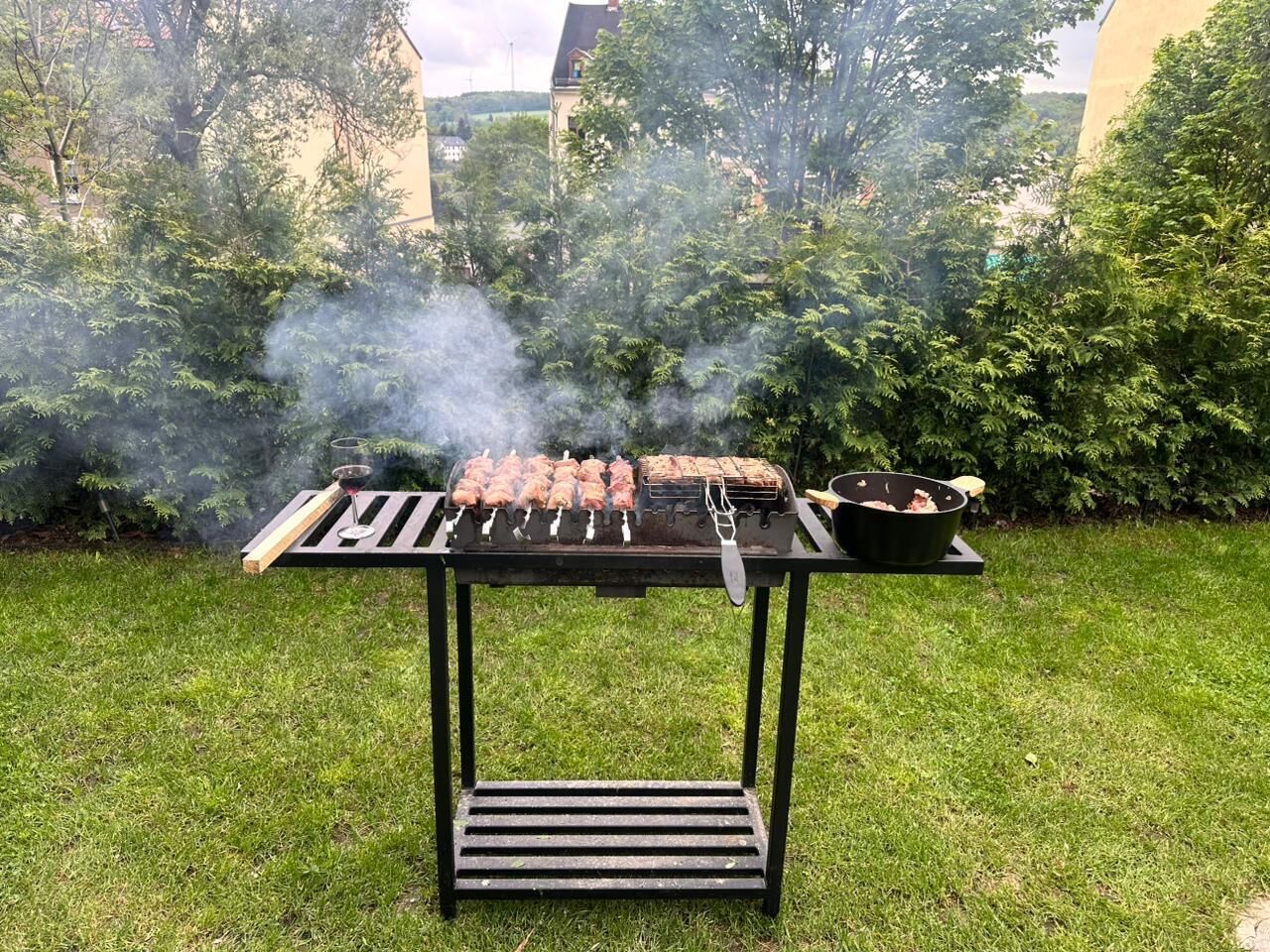 A barbecue grill with skewers of meat, a grill pan, and a smoker, set on a grassy lawn with trees in the background.