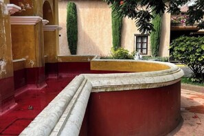 Una panca in marmo curvo con base rossa, una colonna in pietra e un muro con una finestra e vegetazione.