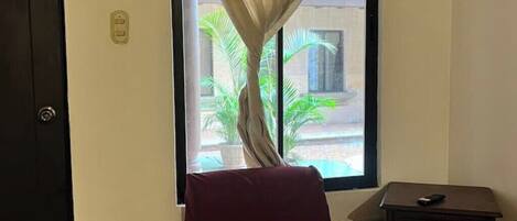 Quarto com cama, janela com cortinas, mesa de cabeceira e vista para palmeiras lá fora.