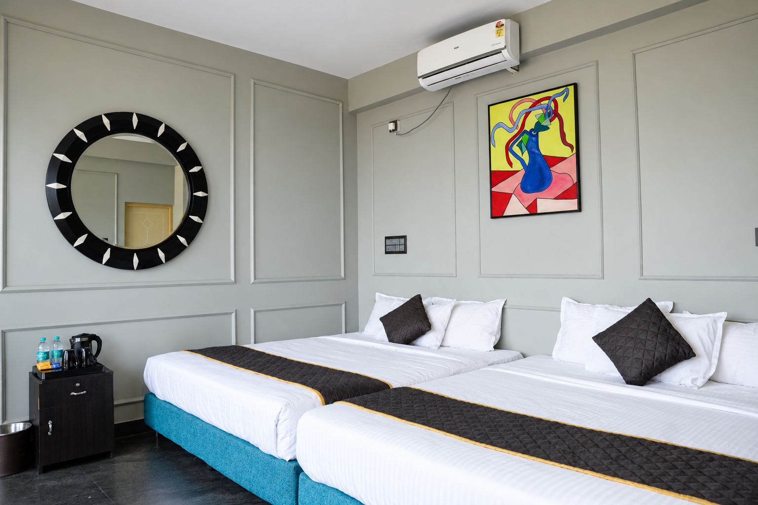 A hotel room with a large bed, a round mirror, an air conditioning unit, and a colorful abstract painting.