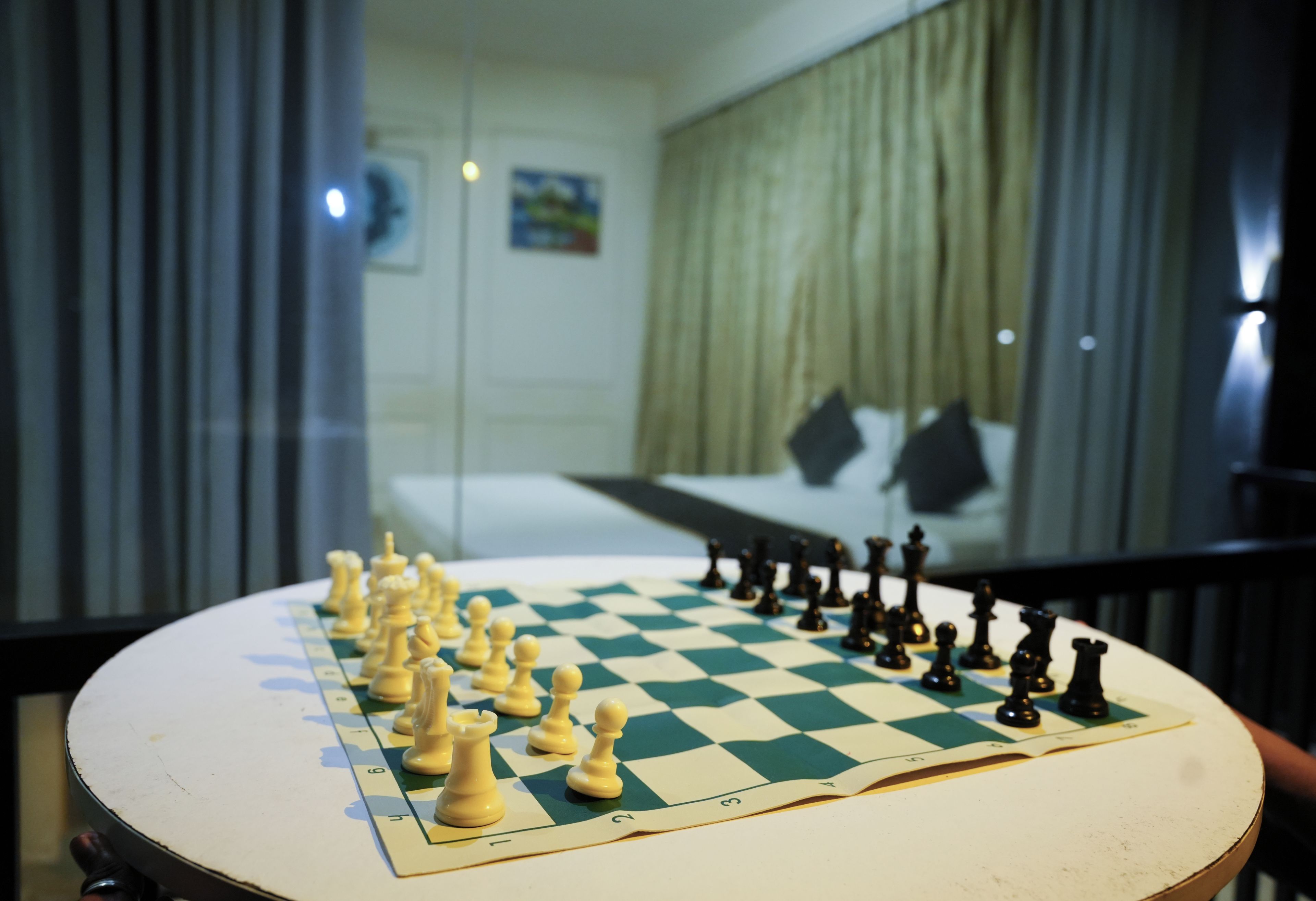 A chessboard with pieces set up in the foreground, a bed with a white headboard and pillows in the background, and a window with curtains.
