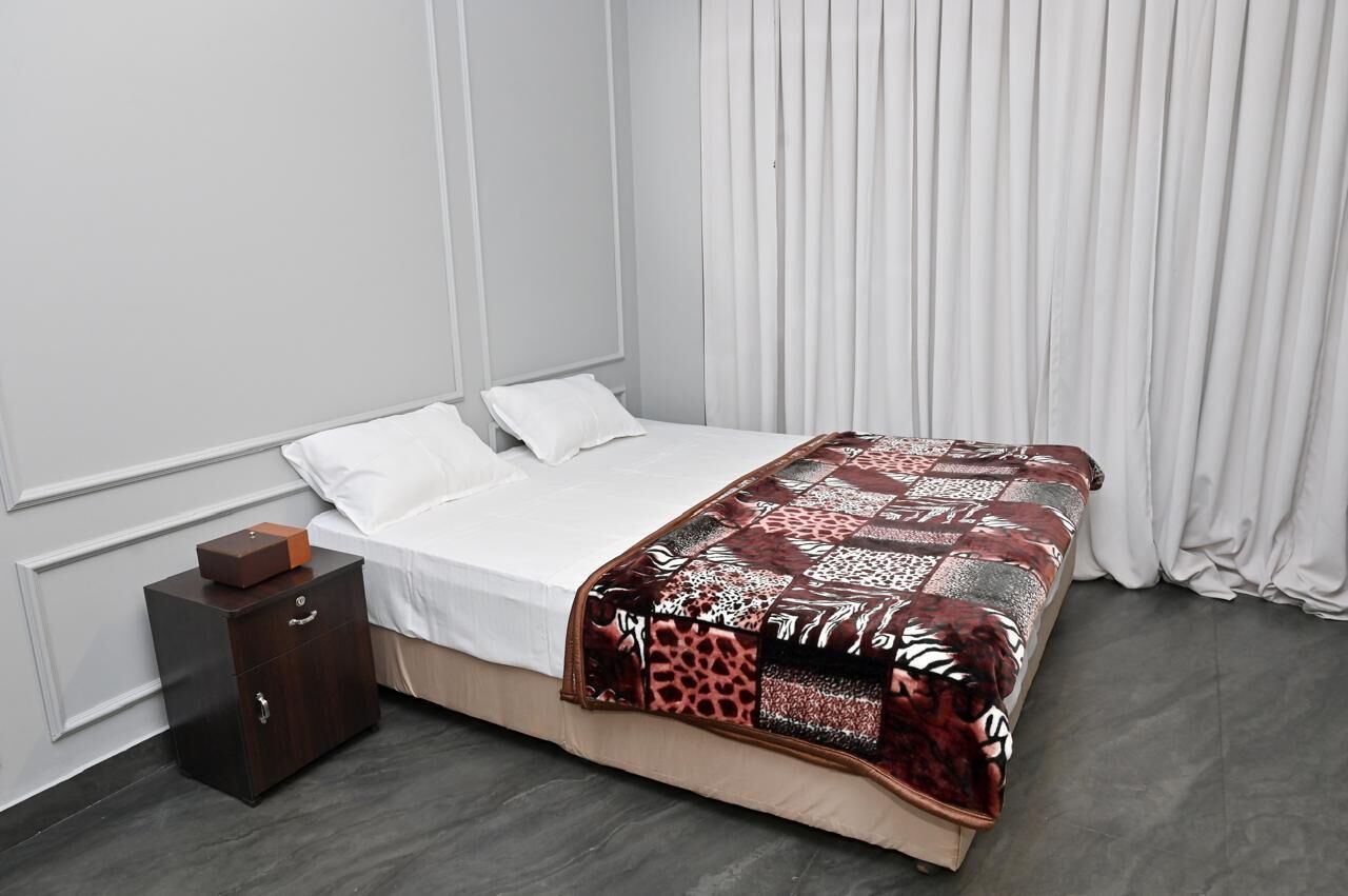 Una cama bien hecha con un edredón estampado, una mesita de noche de madera con cajones y una cortina blanca en una habitación moderna con pared lisa y suelo oscuro.