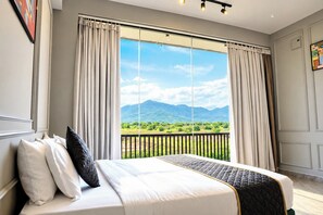 A bedroom with a large bed, two windows with curtains, a view of greenery and mountains, and a balcony with a railing. - GP Golden Palm Meadows Resorts (Bhatkal)