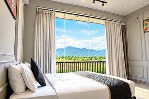 Un dormitorio con una cama grande, dos ventanas con cortinas, vistas a zonas verdes y montañas, y un balcón con barandilla.
