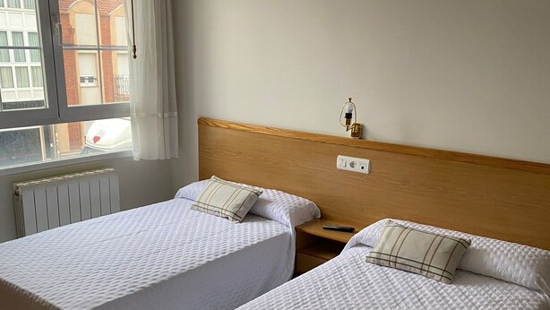 A hotel room with two single beds, a wooden headboard, a bedside table with a lamp, and a window with curtains. - HOSTAL LIBERTAD (Villadangos del Paramo)