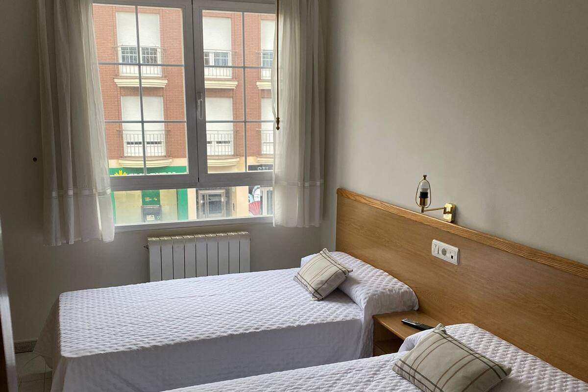 A hotel room with two beds, a window with curtains, a radiator, and a bedside table with a lamp.